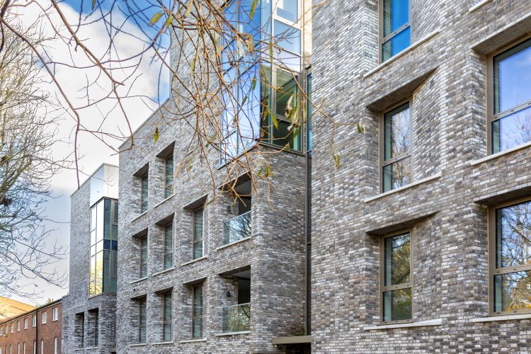 Outside of light-colour brick flats and glass windows reflecting the blue sky and tree, a new development on Elthorne Estate, Islington