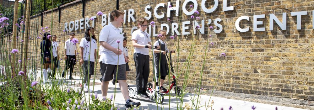 School children walking and using scooters as they walk past a wall with the words 'Robert Blair School and Children's Centre' on it